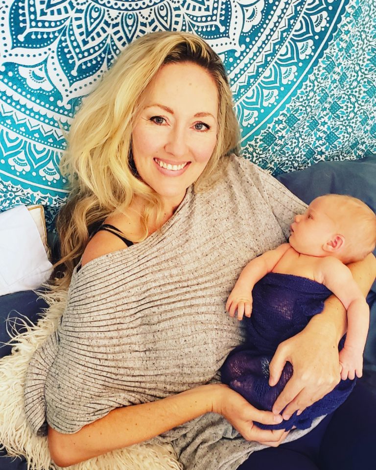 Woman with blond hair wearing a tan poncho holding a baby