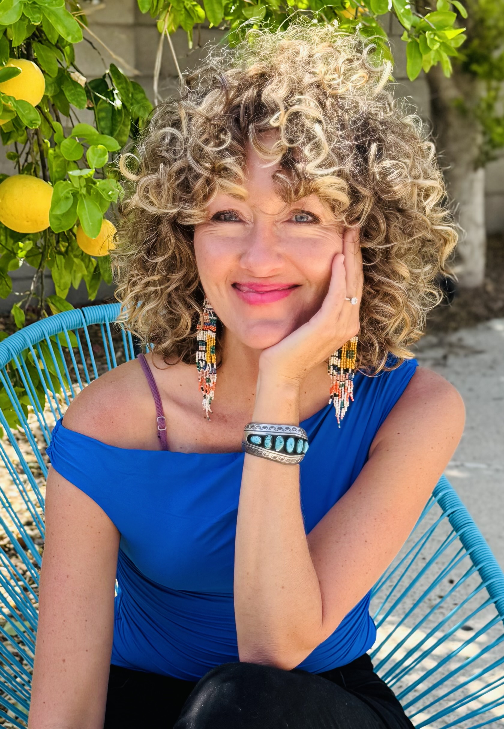 Woman with curly blond hair wearing a blue shirt sitting outside in front of a lemon tree