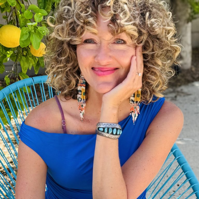 Woman with curly blond hair wearing a blue shirt sitting outside in front of a lemon tree